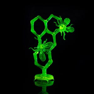 Puede incluir: Una escultura de vidrio verde de un panal con dos abejas. Las abejas están posadas en el panal y tienen alas y cuerpos detallados.