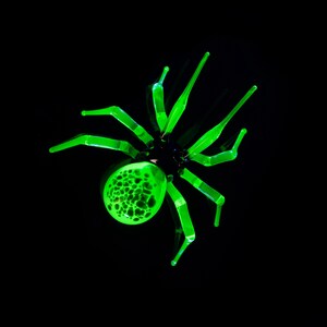 May include: A green glass spider with a textured body and long legs. The spider is set against a black background.