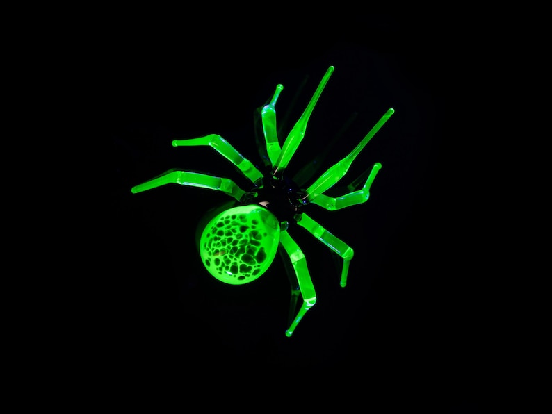 May include: A green glass spider with a textured body and long legs. The spider is set against a black background.