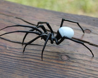 May include: A black glass spider sculpture with a white and black orb-shaped head. The spider is posed with its legs extended, as if it is crawling.