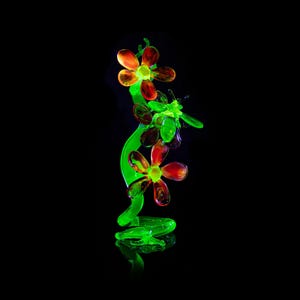 May include: A glass sculpture of a flower stem with three flowers and a bee. The flowers are red and orange, the stem and bee are green. The sculpture is set against a black background.