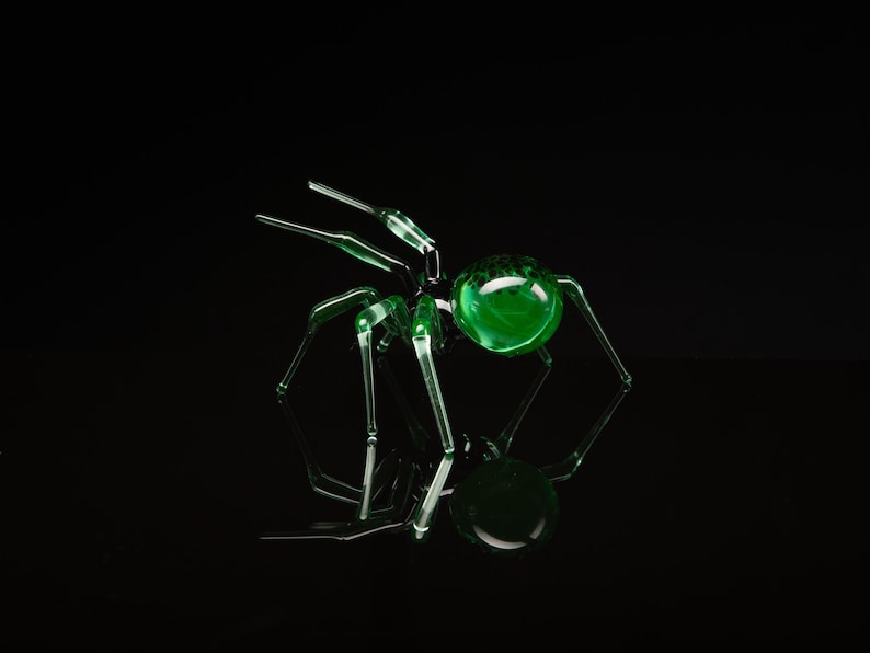 May include: A green glass spider sculpture with a round, green glass body and eight long, thin legs. The spider is sitting on a black surface.