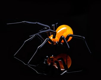 May include: A black glass spider sculpture with an orange glass body. The spider is posed with its legs outstretched and its body tilted forward.