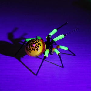 May include: A glass spider sculpture with a black body and green legs. The spider's abdomen is yellow with black spots.