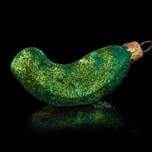 May include: A green, glitter-covered pickle-shaped Christmas bauble. The bauble has a gold-coloured metal cap with a loop for hanging. The pickle is curved and reflects light, creating a sparkling effect against the black background.