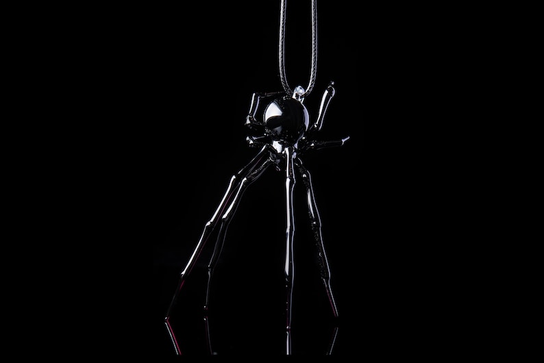May include: A black metal spider pendant necklace with a shiny finish. The spider has eight long legs and a round body.