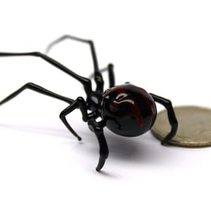 May include: A black spider ornament with a red marking on its back, next to a coin. The spider has long, thin legs and a round body. The coin is silver and provides a size reference.