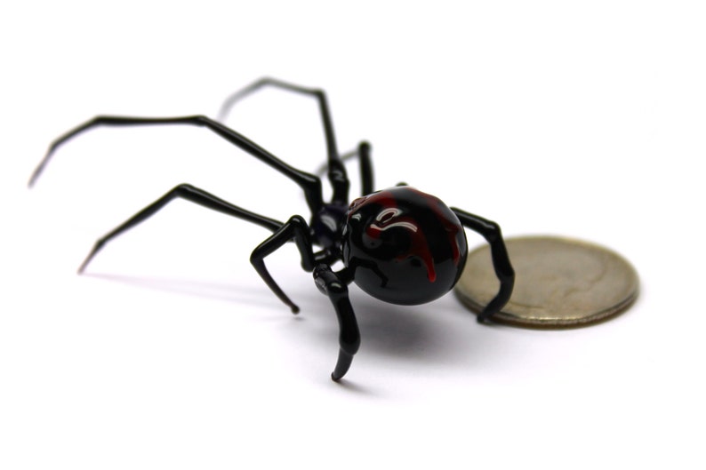 May include: A black spider ornament with a red marking on its back, next to a coin. The spider has long, thin legs and a round body. The coin is silver and provides a size reference.