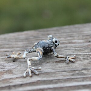 May include: A small, black and white glass frog figurine with detailed features. The frog is posed in a jumping position with its legs outstretched.