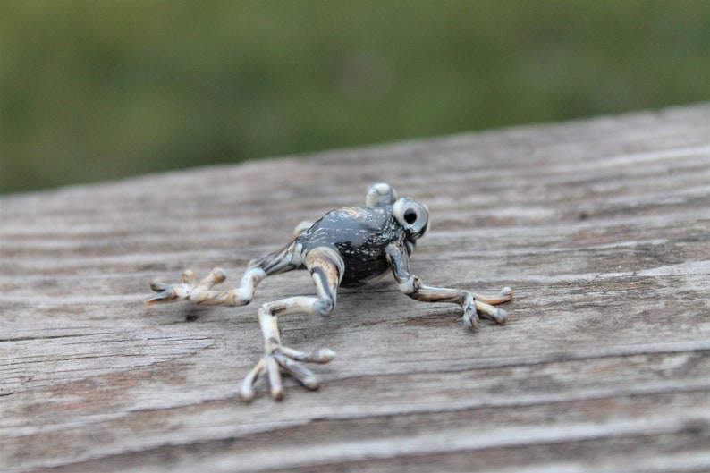 May include: A small, black and white glass frog figurine with detailed features. The frog is posed in a jumping position with its legs outstretched.