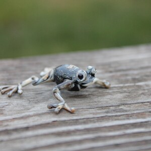 May include: A small, grey and white glass frog figurine with black eyes. The frog is posed in a jumping position with its legs outstretched.