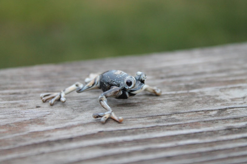May include: A small, grey and white glass frog figurine with black eyes. The frog is posed in a jumping position with its legs outstretched.