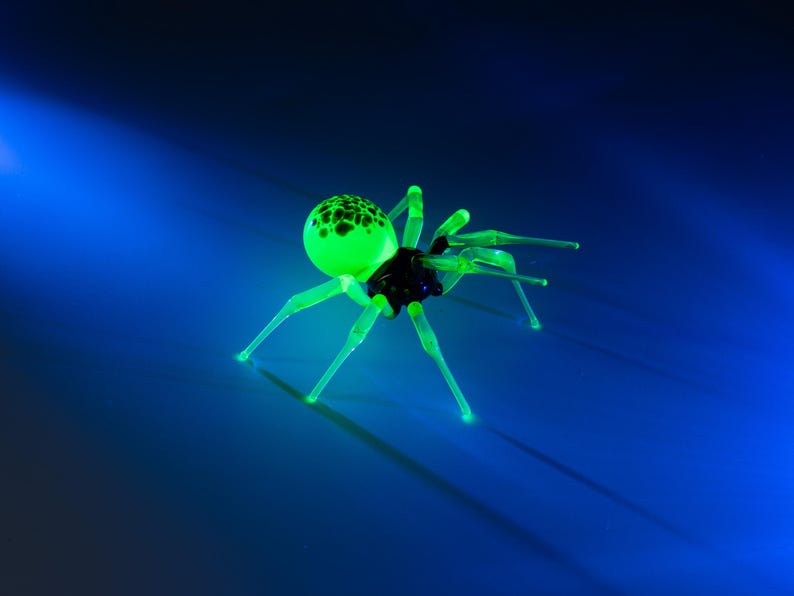 May include: A green glass spider with black spots on its body. The spider has eight legs and is posed in a crawling position. The spider is on a blue background.