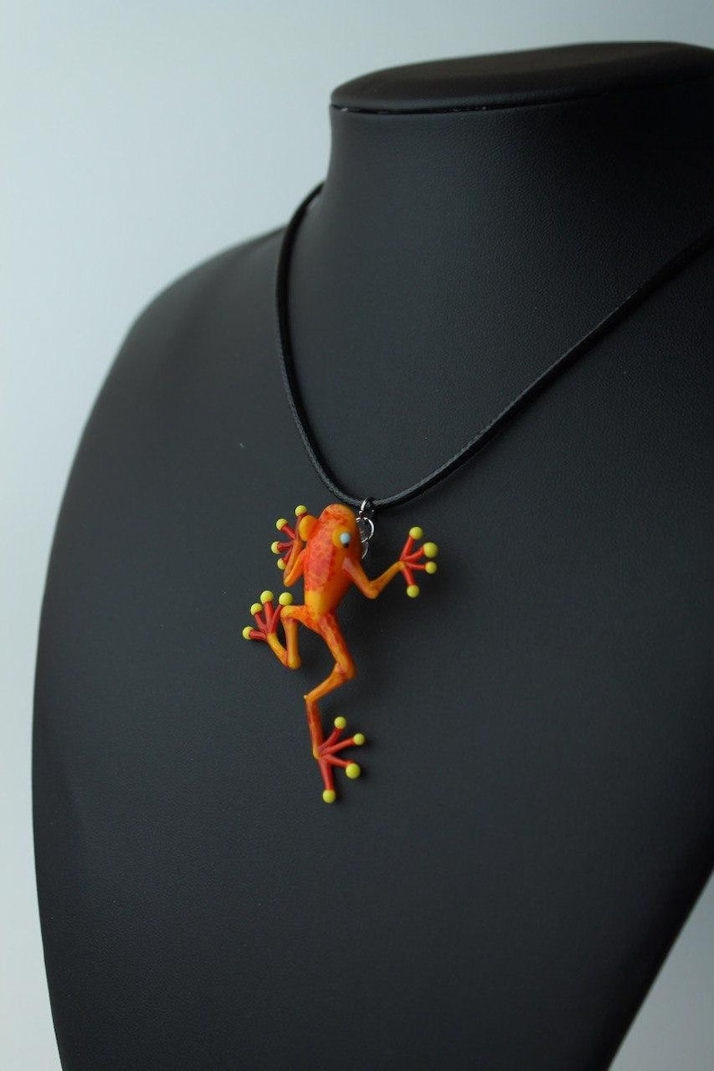 May include: A necklace with a bright orange and yellow frog pendant. The frog is in a climbing pose with its legs outstretched. The necklace is made of black cord.