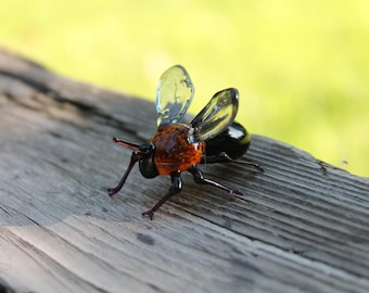 May include: A glass bee figurine with clear wings, a black abdomen, and an orange and brown thorax. The bee is perched on a weathered wooden surface.