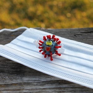 May include: A white face mask with a red and blue cartoon virus figurine on top. The virus is smiling and giving a thumbs up with both hands.