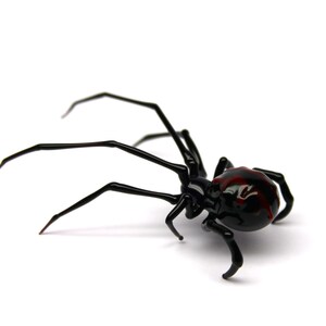 May include: A black spider ornament with a red marking on its back. The spider has long, thin legs and is set against a white background. The ornament is likely made of glass or a similar material.