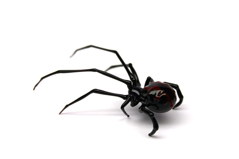 May include: A black spider ornament with a red marking on its back. The spider has long, thin legs and is set against a white background. The ornament is likely made of glass or a similar material.