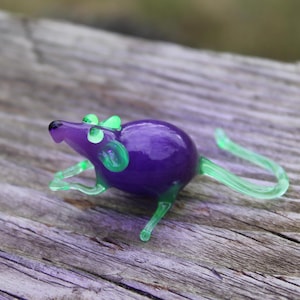 Könnte beinhalten: Kleine Glasmaus-Figur. Die Maus hat einen lila Körper, grüne Ohren, grüne Beine und einen grünen Schwanz. Die Figur liegt auf einer verwitterten Holzoberfläche.