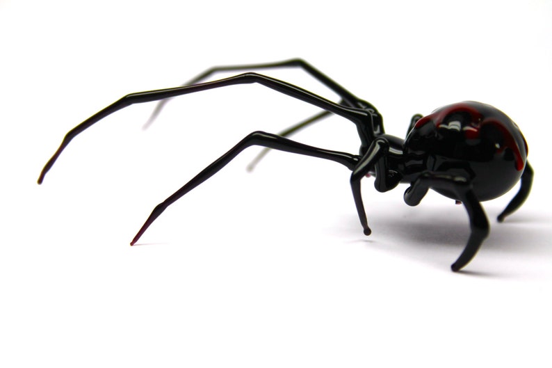 May include: A glossy black spider ornament with long, slender legs and a rounded abdomen. The abdomen has a distinctive red marking. The spider is set against a plain white background, highlighting its sleek, detailed design.