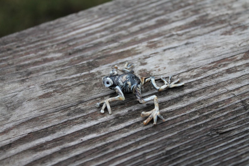 May include: A black and white frog figurine with a detailed design. The frog is posed with its legs outstretched and its eyes are open. The figurine is sitting on a wooden surface.