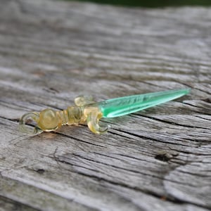 Puede incluir: Una pequeña daga decorativa de vidrio con una hoja verde translúcida y un mango teñido de amarillo. La daga está sobre una superficie de madera gris y desgastada, mostrando sus intrincados detalles.
