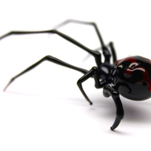 May include: A black spider ornament with long, thin legs and a rounded abdomen. The spider's abdomen has red markings. The ornament is set against a white background.