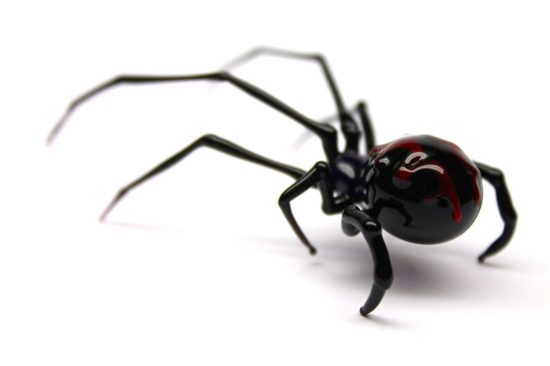 May include: A black spider ornament with long, thin legs and a rounded abdomen. The spider's abdomen has red markings. The ornament is set against a white background.