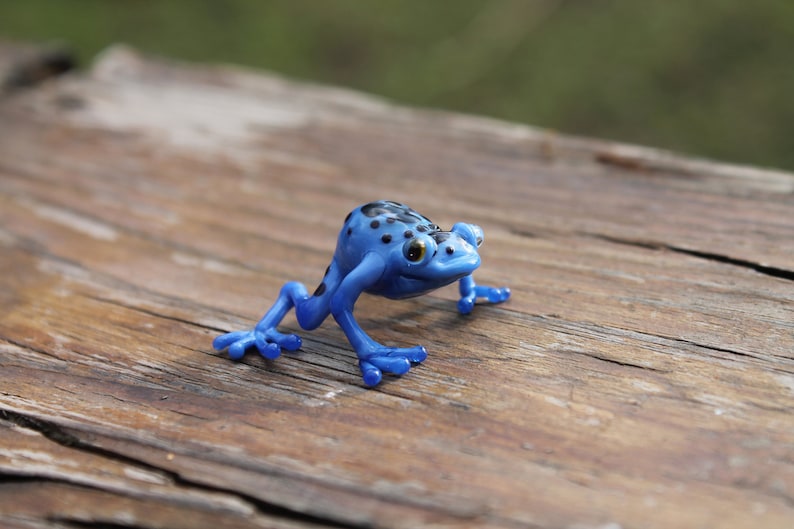 May include: A blue and black spotted frog figurine with black eyes. The frog is posed in a walking position with its legs outstretched. The figurine is sitting on a wooden surface.