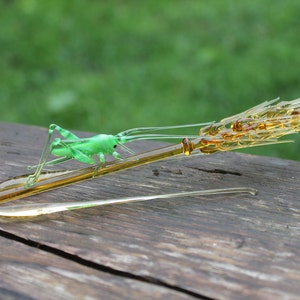 May include: A glass sculpture of a green grasshopper perched on a golden wheat stalk.