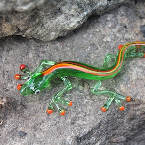 Puede incluir: Una figurita de lagarto de vidrio verde con rayas naranjas y amarillas. El lagarto tiene una cola larga y cuatro patas con dedos naranjas.