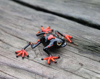 May include: A black glass frog figurine with red and orange accents. The frog has a detailed, realistic design and is posed in a sitting position with its legs outstretched.