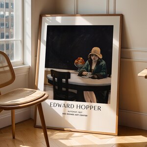 May include: A framed art print of Edward Hopper's "Automat, 1927" from the Des Moines Art Center. The artwork depicts a woman in a hat seated at a table, with a cup and a bowl of fruit. The print is displayed in a room with a window and a wooden chair.