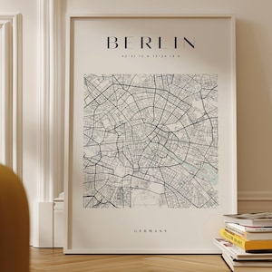 Può includere: Una stampa incorniciata di una mappa di Berlino, Germania. La mappa è in bianco e nero, con il nome della città e le coordinate in alto. La parola "GERMANY" è stampata in basso. La stampa è appoggiata a un muro.