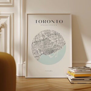 May include: A vertical wall art print featuring a circular black and white street map of Toronto, Canada. The map is set against a cream background with a light blue water section. The text reads TORONTO, the coordinates 43 44 30 N 79 22 54 W, and CANADA.