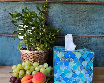 Op de afbeelding: Een blauwe en groene gepatroneerde tissuebox met een wit tissue erop. De tissuebox staat op een houten oppervlak naast een kom met fruit en een potplant.