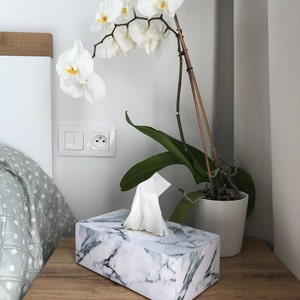 May include: A white marble tissue box with a white tissue dispenser on a wooden nightstand. A white orchid plant with green leaves is in a white ceramic pot on the nightstand.
