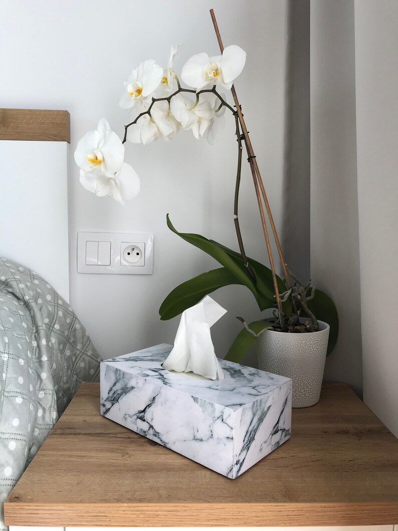 May include: A white marble tissue box with a white tissue dispenser on a wooden nightstand. A white orchid plant with green leaves is in a white ceramic pot on the nightstand.