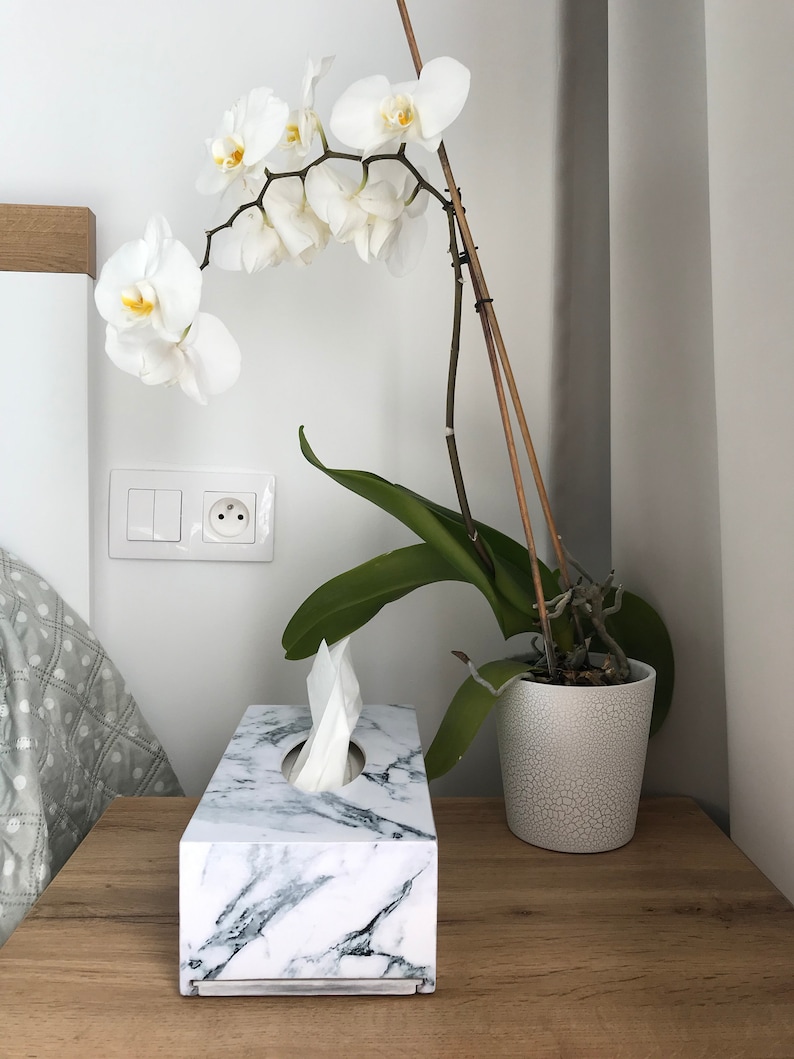 May include: A white marble tissue box holder with a white tissue box inside. A white orchid plant in a white ceramic pot is on a wooden table next to the tissue box.