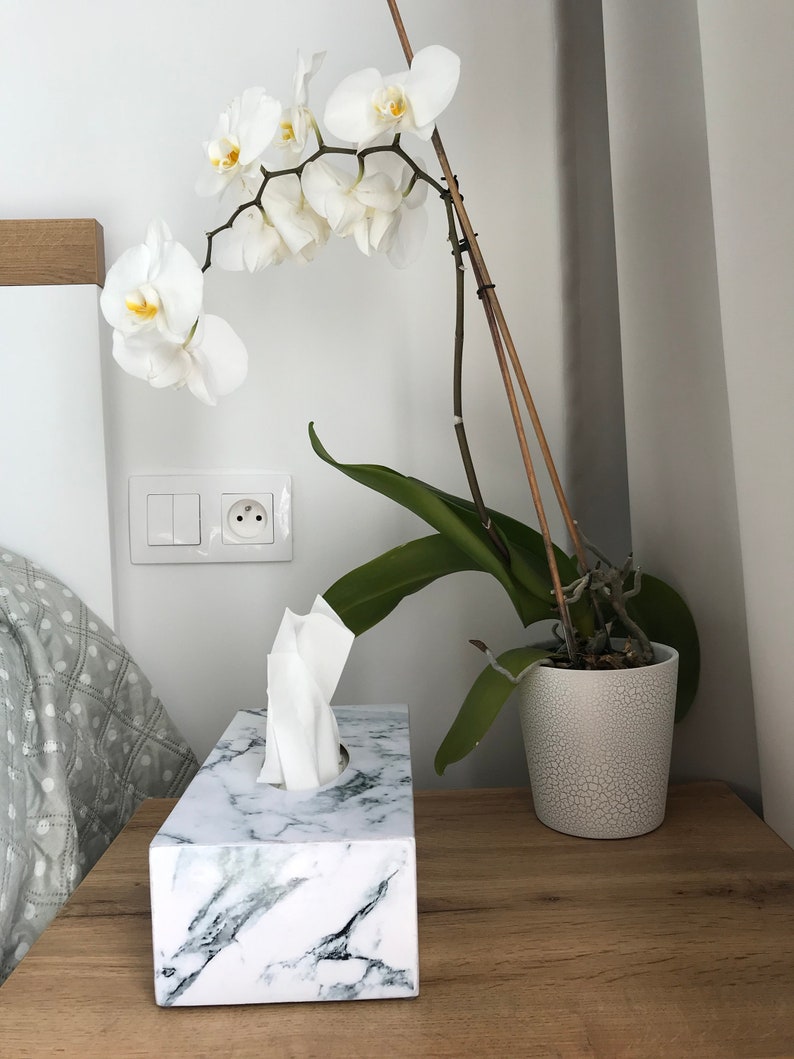 May include: White orchid plant in a white ceramic pot with a marble tissue box holder on a wooden surface.