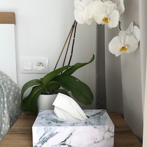 May include: White marble tissue box cover with a white tissue box inside. The tissue box is on a wooden table next to a white orchid plant.
