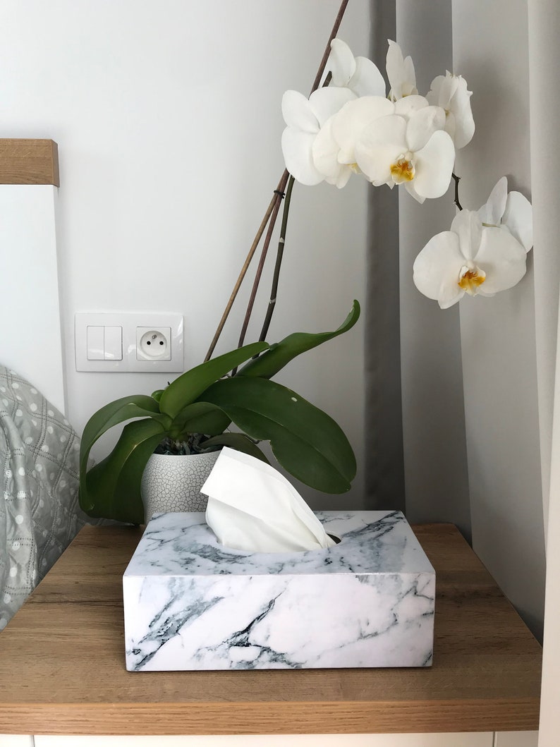 May include: White marble tissue box cover with a white tissue box inside. The tissue box is on a wooden table next to a white orchid plant.