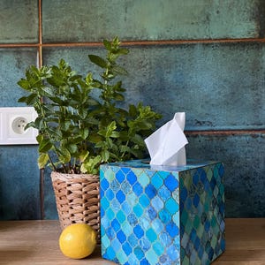 May include: A blue and green tissue box cover with a geometric pattern. The cover is made of wood and has a square shape. The tissue box is sitting on a wooden surface.