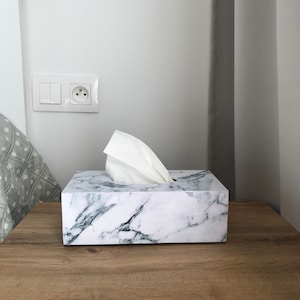 May include: White marble tissue box holder with a white tissue inside. The box is sitting on a wooden surface.