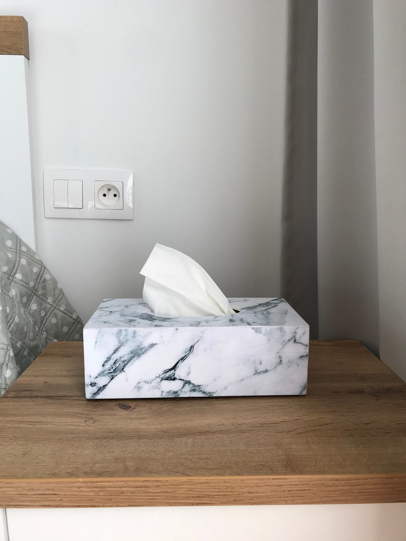 May include: White marble tissue box holder with a white tissue inside. The box is sitting on a wooden surface.