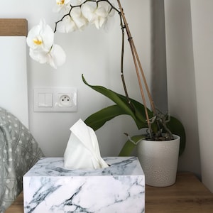 May include: White marble tissue box holder with a white tissue box inside. The tissue box holder is on a wooden surface next to a white pot with a white orchid plant.