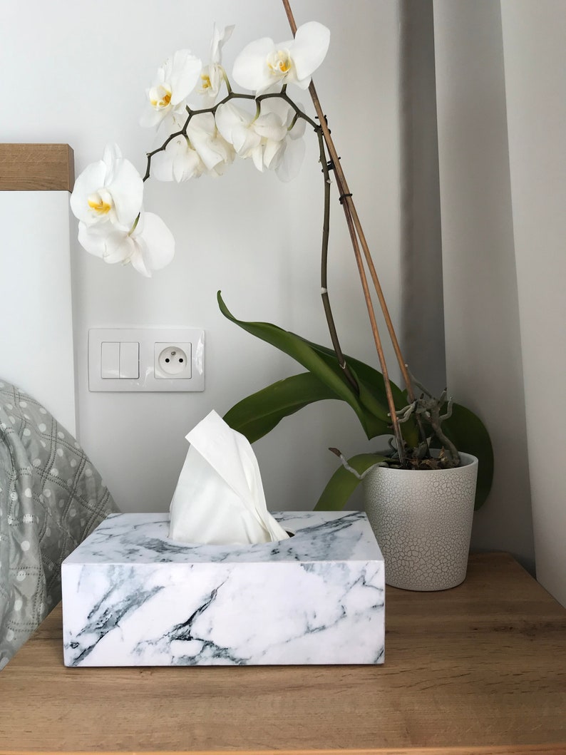 May include: White marble tissue box holder with a white tissue box inside. The tissue box holder is on a wooden surface next to a white pot with a white orchid plant.
