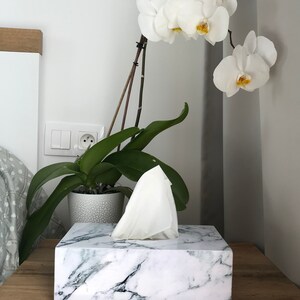 May include: White marble tissue box holder with a white tissue box inside. The tissue box holder is on a wooden surface. A white orchid plant is in the background.
