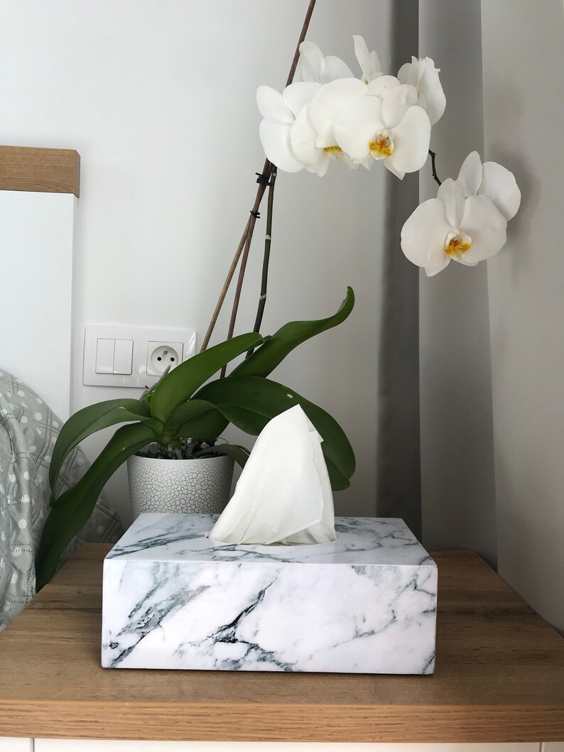 May include: White marble tissue box holder with a white tissue box inside. The tissue box holder is on a wooden surface. A white orchid plant is in the background.