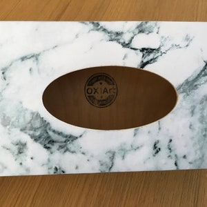 May include: White marble tissue box cover with a rectangular opening for tissues. The cover has a wood-like interior with a black and white logo that says "OXIA&reg;".